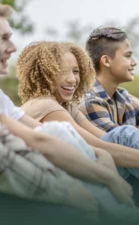 Un groupe de six jeunes est assis côte à côte sur une pelouse, regardant vers le côté droit de l’image et souriant. Ils semblent apprécier la compagnie de l’autre.