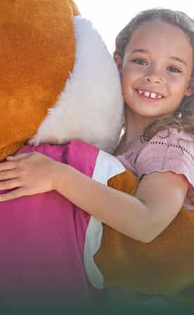 Une petite fille souriante aux cheveux bouclés et vêtue d'un t-shirt rose clair étreint Léa, une grande renarde en peluche brun et blanc.