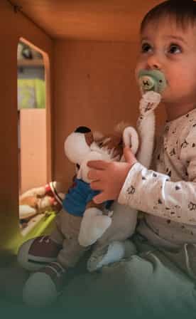 Un jeune enfant avec une sucette dans la bouche est assis et tient une peluche de Léo la mascotte Belambra